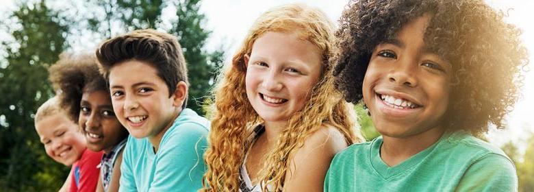 group of racially diverse kids sitting lined up on a bench outside and smiling into the camera